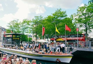 Strommas kanalbåde fyldt med gæster, en rød sightseeingbus ses i baggrunden.