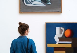 A woman is looking at a painting of a white vase, orange ball, and a book with a blue background. © SMK