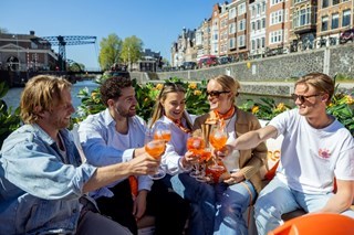 Group of friends sitting on an orange-decorated canal boat toasting with Aperol Spritz glasses while cruising through Amsterdam on a sunny day.