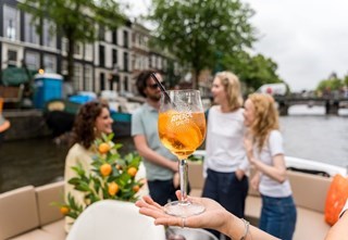 Een glas Aperol Spritz wordt omhooggehouden tegen een achtergrond van vier vrienden die genieten van een gesprek.