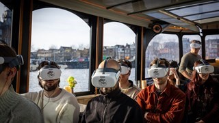 Passagiers dragen VR-headsets in een rondvaartboot tijdens een VR-sightseeingcruise, met de Amsterdamse grachten zichtbaar door de ramen
