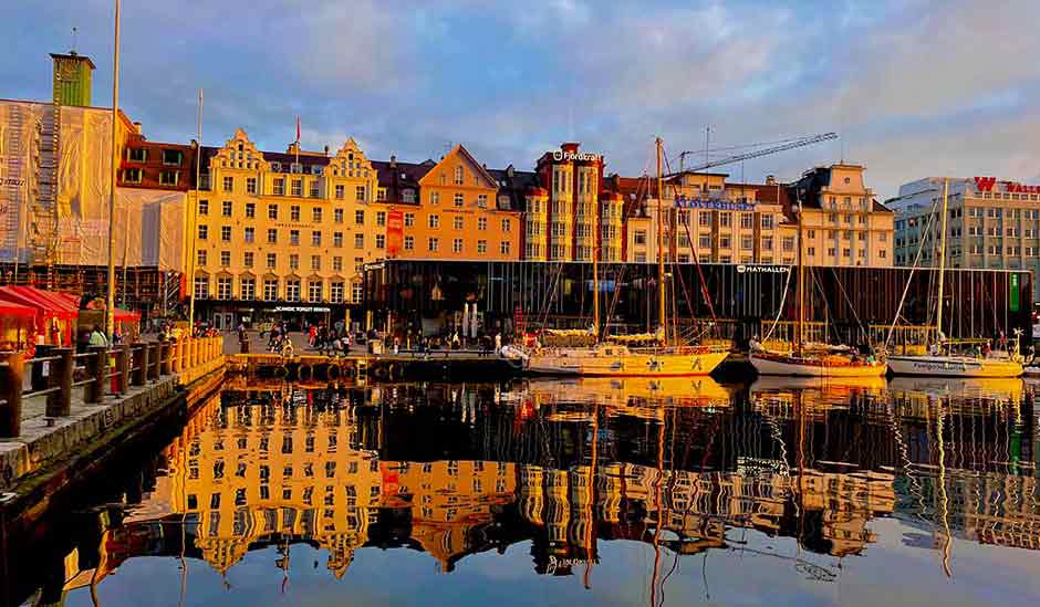 Sailboats moored along the waterfront in Bergen, with historic buildings reflected in the harbor at sunset