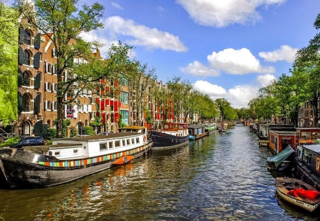 Canal in Amsterdam, Netherlands, lined with houseboats, historic buildings, and trees in the sunshine.