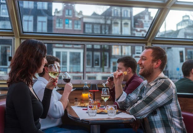 Amsterdam, Niederlande – Gäste genießen Abendessen und Wein auf einer Grachtenfahrt mit Blick auf die Stadt durch das Glasdach