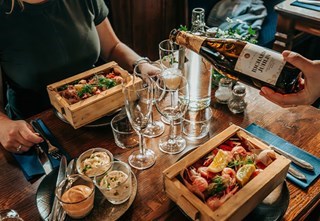  A set table with wooden boxes filled with shrimp, and a guest being served sparkling wine