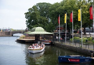 Canal boat departing, with flags and another boat in the background.