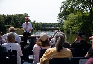 Tour guide speaks to passengers on a canal boat, with greenery in the background.