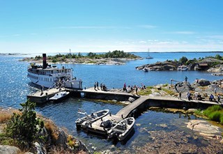 Waxholm III vid bryggan på Bullerö i skärgården, med passagerare, klippor och båtar under en klarblå himmel.