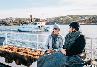 Passengers aboard M/S Angantyr enjoying the winter sun under warm blankets with coffee – another archipelago boat passing behind.