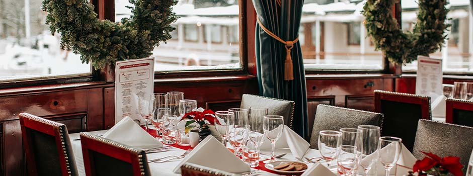 Christmas-decorated dining room onboard a boat - wreaths of pine branches in the windows and festive tables set for the holiday.