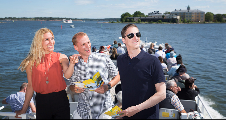Three smiling persons onboard a sightseeing boat. The Suomenlinna Sea Fortress can be seen in the background.